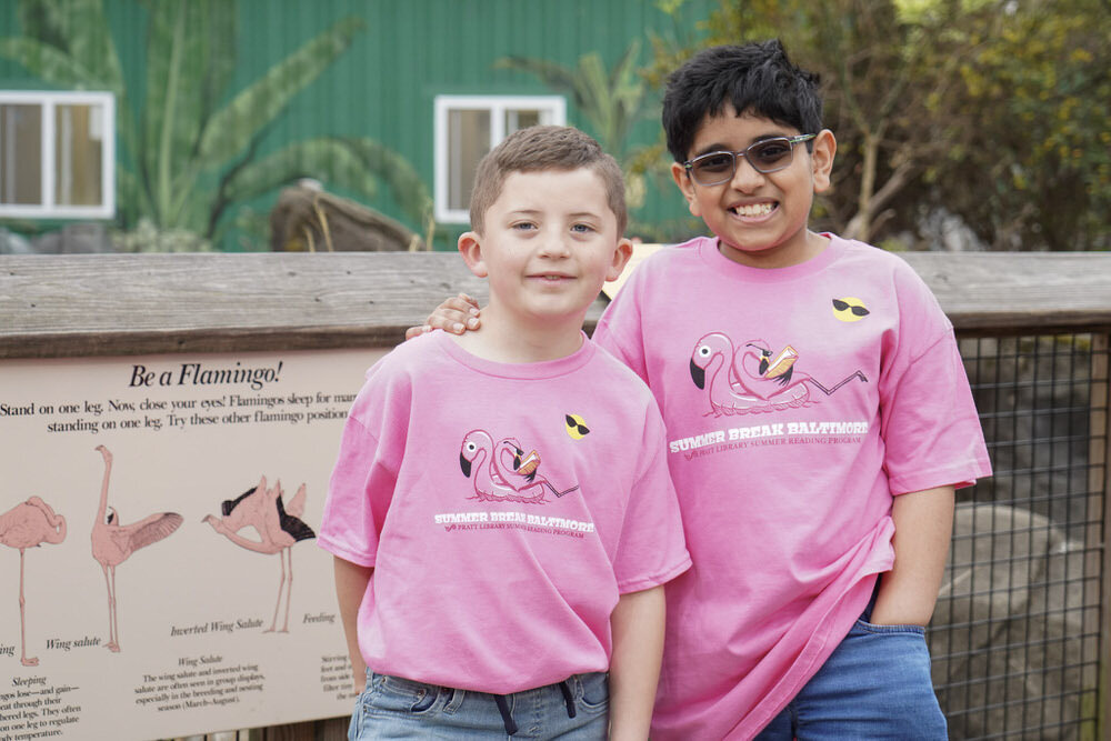 two kids in Summer Break Baltimore t-shirts by a zoo sign reading Be A Flamingo!