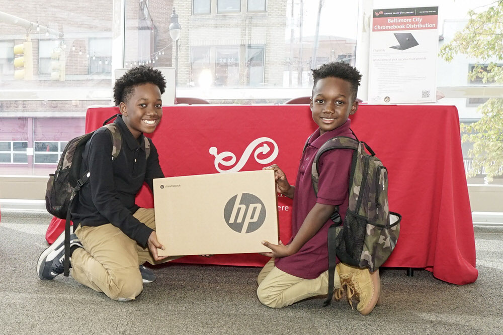 Chromebook distribution - smiling kids with boxed Chromebook by a Pratt Library table